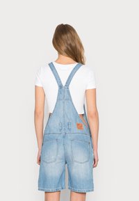 Ljusblå denimoverall med en figurnära crop top. Har justerbara axelband, två bakfickor och en brun läderimitation logotyp patch.