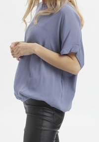 Femme portant un haut lavande à manches courtes et amples en tissu texturé, associé à un pantalon en cuir noir, se tenant devant un fond blanc uni.