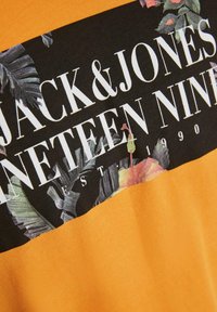 Tessuto arancione con una toppa rettangolare nera che presenta il testo bianco "JACK & JONES NINETEEN NINETY" e design floreali.