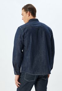 Mörk denimskjorta med krage, långa ärmar och knappmanchetter. Har kontrasterande stygn och rak fåll. Texturen är slät.