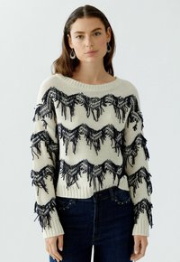 Cremefarbener Strickpullover mit marineblauen Fransenakzenten und Wellenmustern. Hat einen weiten Ausschnitt und gerippte Bündchen, kombiniert mit dunklen Jeans.