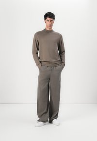 Maglione a maglia marrone cioccolato con collo alto, abbinato a pantaloni grigi a gamba larga. Scarpe da ginnastica bianche completano l'outfit.