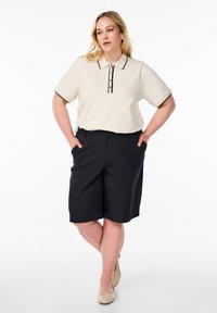 Svetlo béžové polo tričko s krátkym rukávom, čiernymi okrajmi a gombíkovou légou, spárované s čiernymi šortkami po kolená. Model stojí s rukami v vreckách.