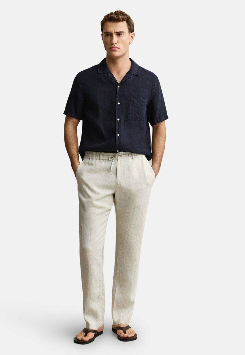 Man draagt een donkerblauw overhemd met korte mouwen en knopen, licht beige broek met trekkoord en bruine sandalen, staand met handen in de zakken.