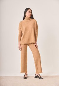 Suéter de punto color tostado con cuello alto y dobladillo con flecos, combinado con pantalones de pierna ancha a juego. El modelo lleva zapatos de charol negros.