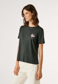 T-shirt a maniche corte verde scuro in tessuto morbido, con una stampa grafica rosa sul petto e elementi testuali, vestibilità casual e scollo rotondo.