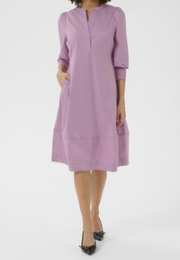 Fioletowa sukienka do kolan z kołnierzykiem, rękawami o długości trzech kwart, kieszeniami i subtelnymi fałdami. Gładka faktura i luźny krój. Czarne, spiczaste buty.