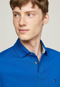 Niebieska koszulka polo z teksturowanej tkaniny, z kołnierzykiem, dwoma guzikami i małą naszywką z logo.
