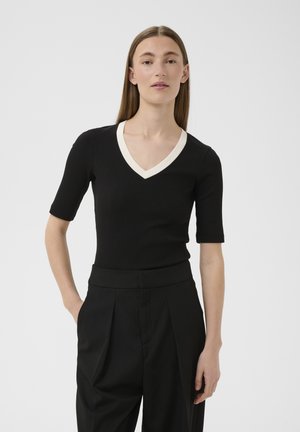 Frau mit glattem braunem Haar, die ein schwarzes V-Ausschnitt-Kurzarmshirt mit weißem Besatz und hoch taillierte schwarze Hose vor einfachem Hintergrund trägt.