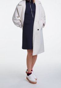 Weißer, strukturierter Mantel über einem marineblauen Kleid mit einem Reißverschluss auf der Vorderseite und einer Tasche. Abgerundet mit weißen Sneakern und roten Socken an den Knöcheln.