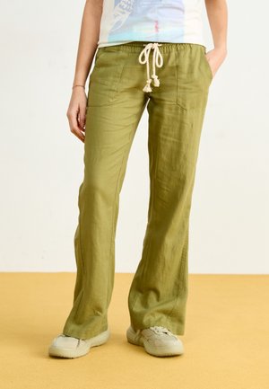 Personne portant un pantalon vert olive à cordon avec poches avant et des baskets blanches, debout sur un sol beige.