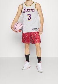 Maglia dei Lakers grigia con dettagli viola, pantaloni corti rossi a motivi, calze nere e sneakers bianche, mentre tiene un pallone da basket rosa.
