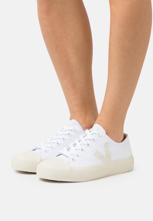 Baskets en toile blanches avec une semelle en caoutchouc, arborant un logo beige clair sur le côté, bout arrondi et six œillets pour les lacets.