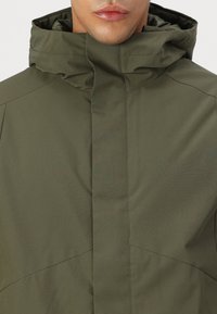 Olivegrønn jakke laget av glatt stoff, med høy krage og minimalistisk design med skjulte lukkinger og subtile sømdetaljer.