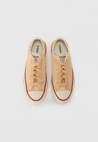 Zapatillas de lona beige con puntera de goma blanca, franja roja y seis ojales de metal. Los cordones son beige y el forro interior muestra el nombre de la marca.