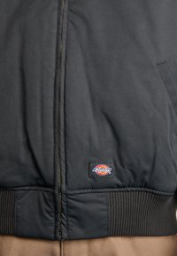 Jachetă bomber neagră cu fermoar, manșete ribbed și logo cu patch. Materialul pare neted, cu un finisaj mat.