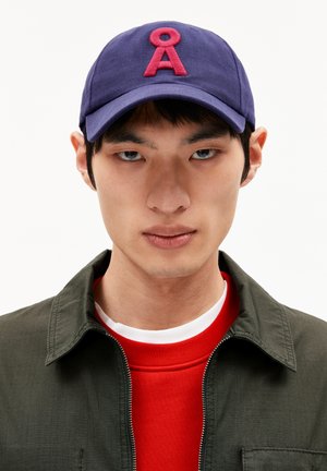 Purple cap with a raised pink "A" logo on the front. The wearer has a green jacket and a red sweatshirt with a white neckline.