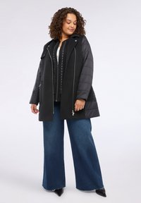Abrigo negro con capas y sección superior acolchada, detalle de cremallera y bolsillos laterales. Combinado con pantalones de mezclilla azul de pierna ancha.