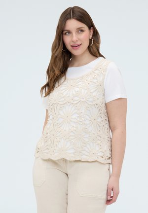 Kvinde med langt brunt hår iført øreringe i ringform, en hvid top med korte ærmer og en beige blomsterhæklet ærmeløs overdel.