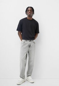 Maglietta nera a maniche corte, jeans chiari strappati, e sneakers bianche. Il modello sta in piedi con le mani dietro, su uno sfondo neutro.