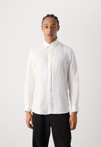 Chemise en lin blanche avec un col, des manches longues et des boutons à l'avant. Texture douce avec un aspect légèrement froissé. Portée avec un pantalon noir.