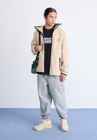 Chaqueta de forro polar beige con detalles en negro, camiseta gráfica negra, pantalones jogger grises y zapatillas ligeras. Un bolso cruzado verde añade un toque de color.