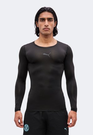 Ung mand med mørkt hår iført en tætsiddende sort langærmet Puma trøje og sorte shorts med blå logoer, står foran en ensfarvet baggrund.