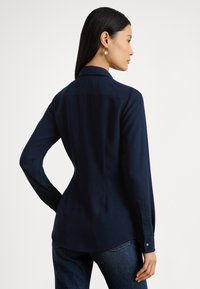 Tumši zils krekls ar garām piedurknēm, apkakli, pogātas aproces, aizmugures šuves detaļu un gludu tekstūru. Kombinēts ar tumšiem džinsa biksēm.