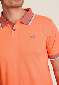 Oranje polo shirt met een gestructureerde kraag met een rood en blauw geruit patroon, een klein logo-opdruk op de borst en contrasterende afwerking.