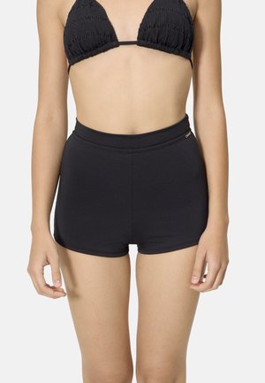 Mujer con top de bikini negro texturizado y pantalones cortos de baño negros de tiro alto, de pie con los brazos relajados a los lados.