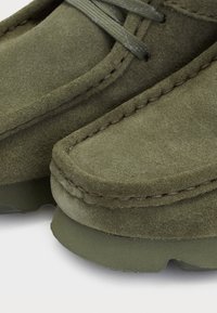 Chaussures en daim vert avec des bouts ronds, des coutures visibles et des détails en lacets. La semelle est texturée avec une apparence plate et durable.
