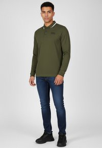 Ung man står iförd en olivgrön långärmad polo-shirt, blå slim-fit jeans och svarta sneakers mot en enfärgad bakgrund.