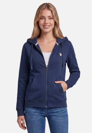 Smiling woman wearing a navy blue zip-up hoodie with a small logo on the chest, white top underneath, and blue jeans, standing with hand in pocket.