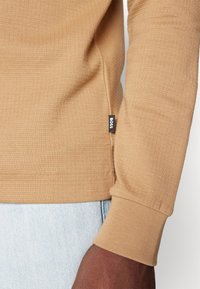 Pullover beige a maniche lunghe realizzato in tessuto strutturato, con polsini aderenti e una piccola etichetta nera sul lato.