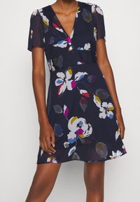 Blumenkleid in Marineblau mit kurzen Ärmeln, V-Ausschnitt und lockerer Passform. Es hat bunte Blumenmuster in Pink, Gelb und Blau. Weicher Stoff.