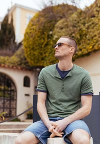 Green short-sleeve henley shirt with a navy inner collar, paired with denim shorts. The person wears dark sunglasses and sits outdoors.