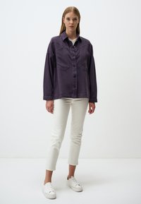 Camicia oversize viola con due tasche sul petto, abbinata a jeans skinny bianchi e sneakers bianche, indossata da un modello su uno sfondo neutro.