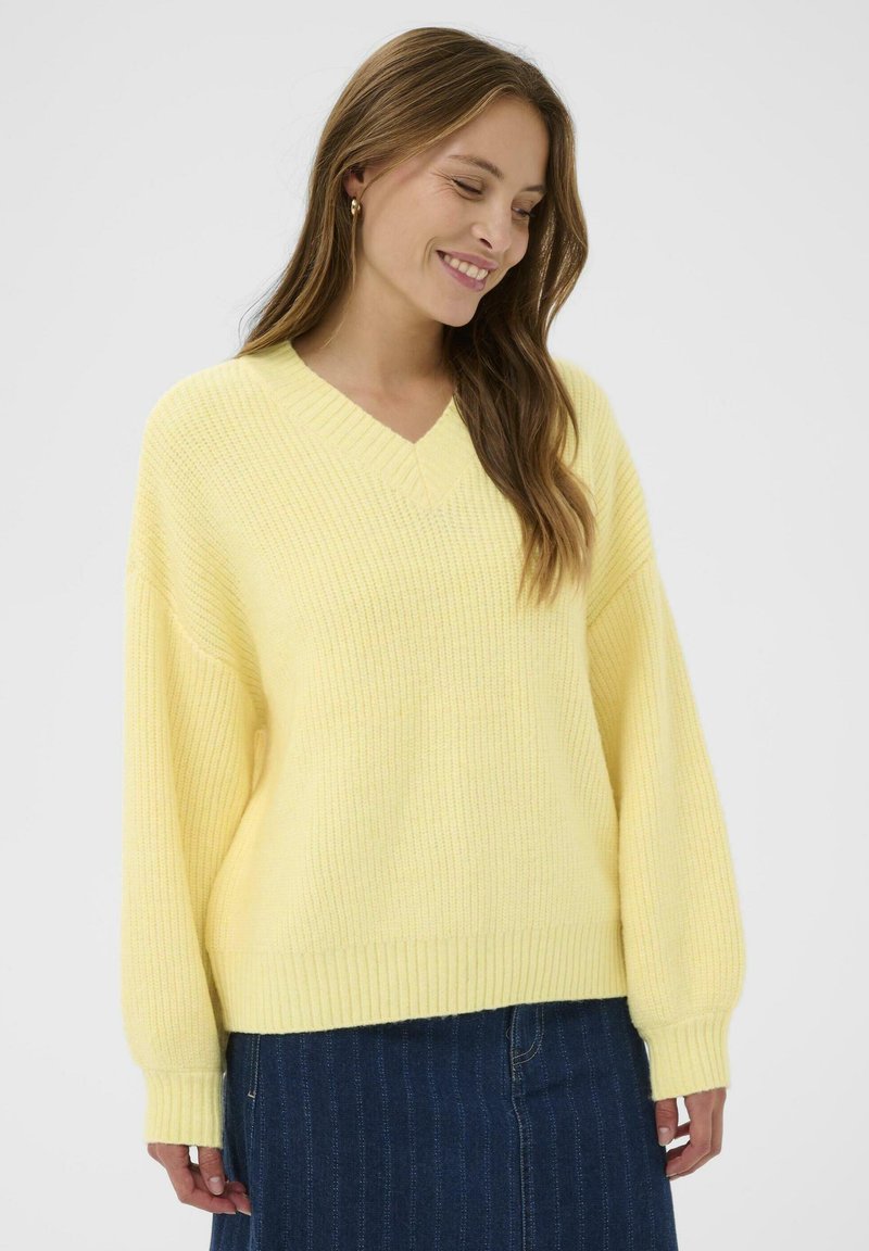 Pull jaune en tricot avec un col en V, épaules tombantes et ourlet côtelé. Texture douce, coupe décontractée, associé à une jupe en denim.