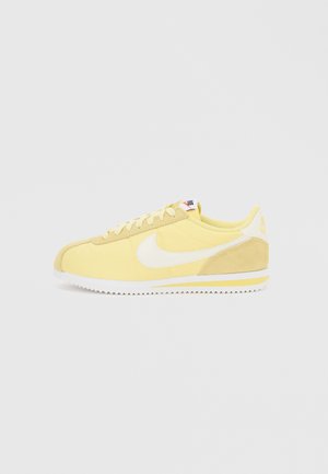 Zapatilla Nike Cortez amarilla con el logo Swoosh blanco, detalles en gamuza beige, suela blanca con zigzag y entresuela amarilla con rayas, vista lateral.