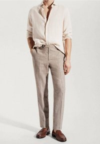 Mężczyzna w jasnobeżowej lnianej koszuli, włożonej w jasnobrązowe dopasowane spodnie, w brązowych skórzanych mokasynach, stojący z jedną ręką w kieszeni.