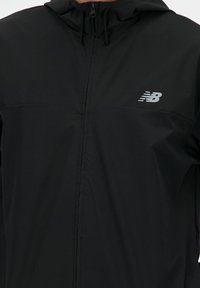 Giacca nera con zip e cappuccio, realizzata in tessuto morbido. Logo New Balance di colore grigio in evidenza sull'area superiore sinistra del petto.