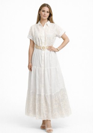 Femme portant une robe maxi blanche à manches courtes boutonnée avec broderie florale, ceinture tissée beige, montre, bracelet et talons ouverts couleur nude.