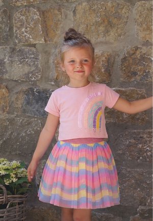 Junges Mädchen mit Dutt-Frisur trägt ein rosa Hemd mit einem Regenbogendesign und einen pastellgestreiften Rock, steht vor einer Steinmauer.