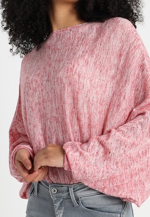 Vrouw draagt een losse, lange mouwen roze gespikkelde trui, ingestopt in lichtgrijze spijkerbroek, haar handen zachtjes samengevouwen.