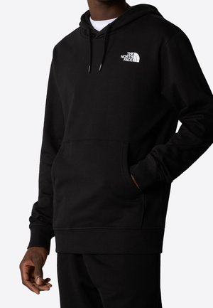 Mann trägt schwarzen The North Face Hoodie mit Fronttasche und weißem Logo auf der Brust, vor einfachem hellem Hintergrund.