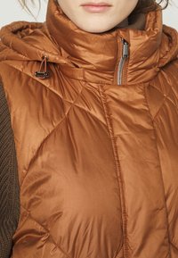 Tikkitettu, hihaton ruskea toppaliivi, jossa korkea kaulus. Valmistettu kiiltävästä, sileästä kankaasta; edessä vetoketju ja kiristysnyörit.