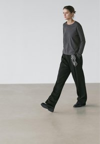 Femme marchant à l'intérieur, portant une chemise à manches longues gris foncé, un pantalon large noir avec une écharpe, et des chaussures noires sur un sol lisse.