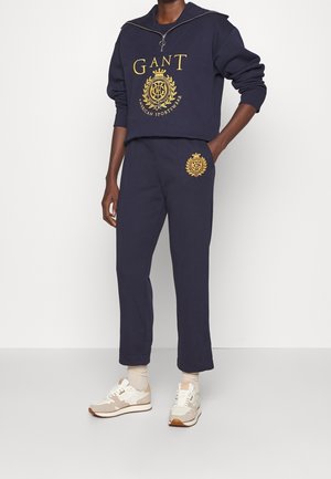 Mężczyzna w granatowym dresie GANT z żółtym logo na piersi i udzie, połączonym z beżowymi i białymi sneakersami oraz jasnymi skarpetami.