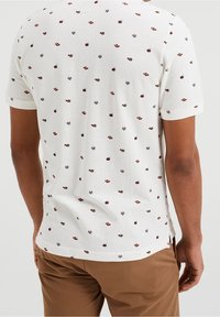 T-košile s krátkým rukávem v krémové barvě, vyrobená z texturované látky, se rozptýleným květinovým vzorem v červené, modré a zelené barvě.