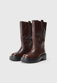 Botas de cuero marrón hasta la rodilla con una suela negra gruesa, textura suave y tiradores laterales para facilitar su uso. Costuras visibles en la parte superior.